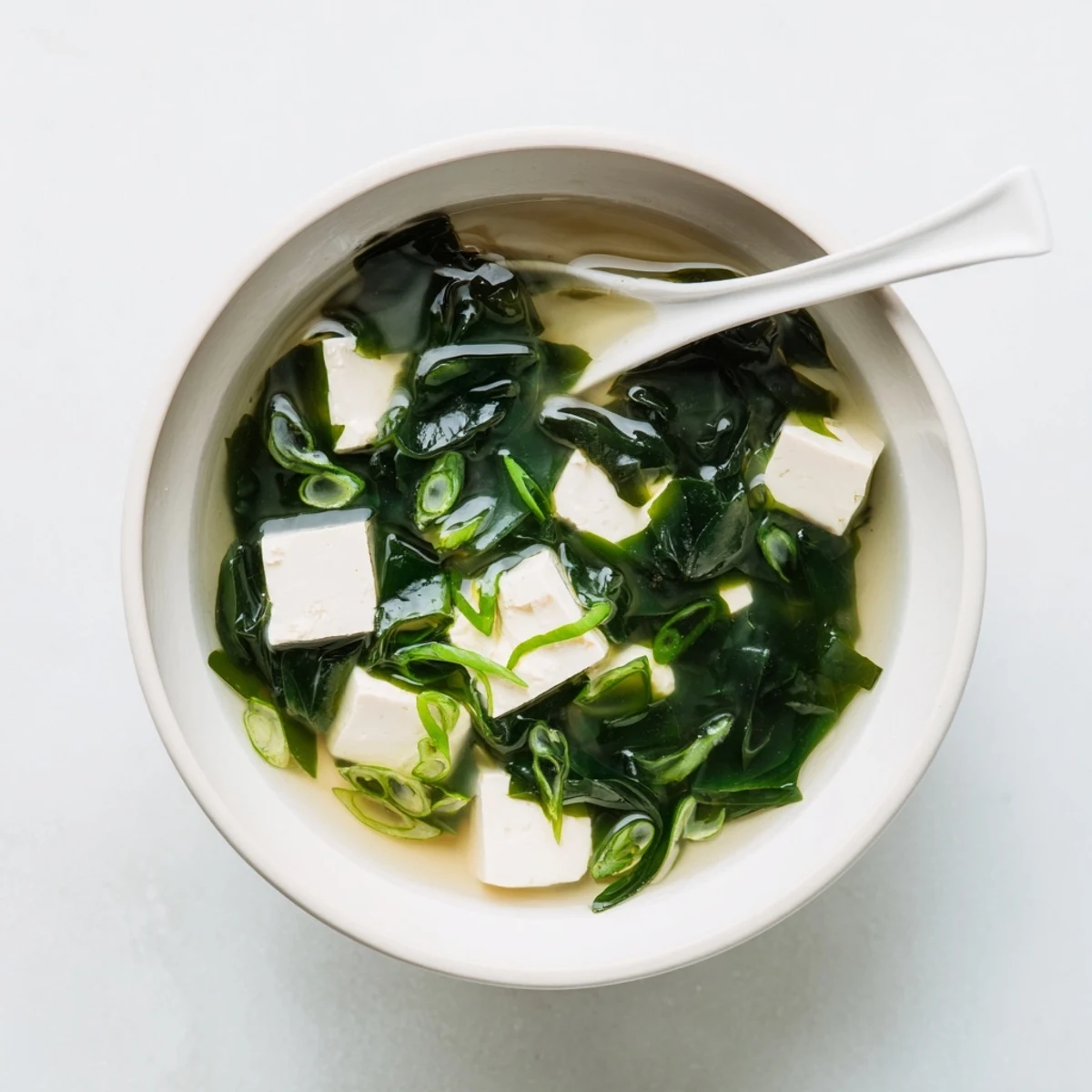 A close-up of steaming wakame soup, highlighting tender seaweed, soft tofu cubes, and vibrant green scallions.